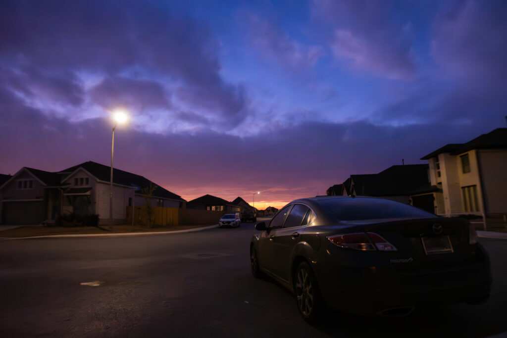 A low-light photograph capturing the serene ambiance of a suburban neighborhood in the hill country area of San Antonio, Texas, as dusk settles in, highlighting the gentle illumination of street and house lights against the soft gradients of the evening sky.